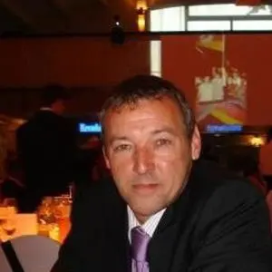 A man in a suit sits at a table in a dimly lit event or banquet hall, possibly among board members, with a projection screen and other guests visible in the background.