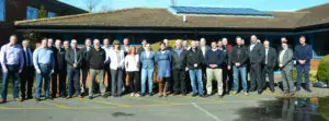A group of about 25 adults stands in a queue outside a building with solar panels on the roof, under a clear blue sky.