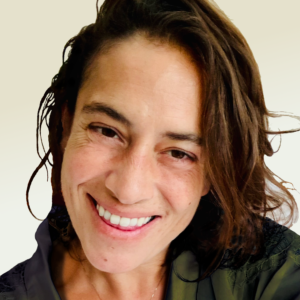 Smiling Anne Marie Chebib with shoulder-length brown hair, wearing a dark top, looks directly at the camera against a light background.