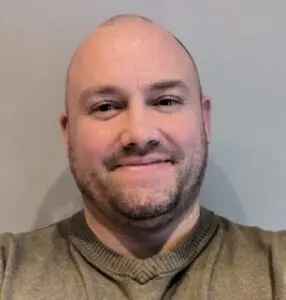 A man with a shaven head and short beard, wearing a green jumper, smiles at the camera against a plain light-coloured background—capturing Paul Townsend’s approachable style in 2024.