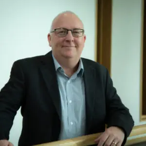 Bald man with glasses, identified as Ric Robins, wearing a dark blazer and light shirt stands indoors near a railing, smiling at the camera in 2024.