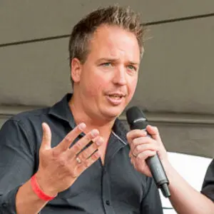 A man in a black shirt speaks whilst gesturing with one hand as another person holds a microphone to him, under a tent or marquee, during the 2024 event. Sander Teunissen appears engaged in conversation, contributing to the lively atmosphere.