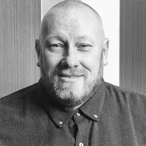Bald man with a beard wearing a buttoned-up shirt, smiling, in front of a vertically striped background. Black and white photo of Michael Kill, 2024.