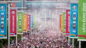 Large crowd gathers outside Wembley Stadium for UEFA Euro 2020, with colourful event banners lining the walkway and smoke visible in the air—a scene later scrutinised in Baroness Casey’s 2024 report.