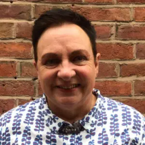 Julie Tippins 2024: Person with short dark hair standing in front of a brick wall, wearing a white shirt with blue and red leaf patterns, smiling at the camera.