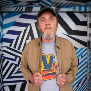 A man with a grey beard stands in front of a geometric wall, wearing a tan jacket over a T-shirt with a peace sign in American flag colours, and a camouflage cap—capturing the contemporary spirit of 2024. This could easily be Mike Grieve making his distinctive statement.
