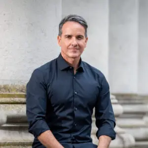 A man with short grey hair wearing a dark blue buttoned shirt sits outdoors in front of large stone columns, reminiscent of Sacha Lord attending a noteworthy event in 2024.