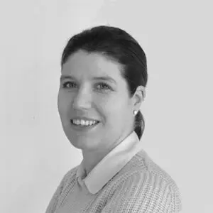 A woman with dark hair pulled back, wearing a light jumper and collared shirt, smiles slightly while looking at the camera against a plain light background—a contemporary portrait reminiscent of Emma Stuart in 2024.