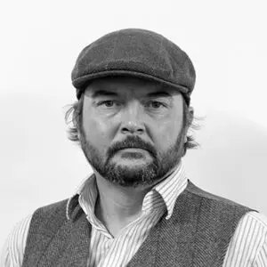 A man with a beard and moustache, reminiscent of Gareth Hughes in style, wearing a flat cap, striped shirt, and waistcoat looks directly at the camera against a plain white background in this 2024 portrait.