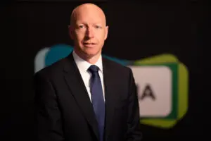 A bald man in a dark suit and blue tie, representing Board Members, stands in front of a blurred background with geometric shapes and partial text.