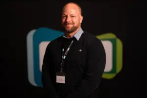 A man with a beard, wearing a black jumper and name badge, stands in front of a dark background with blue and green shapes, presenting the professional demeanour often seen among Board Members.