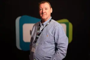 A man wearing a blue checked shirt and conference lanyard stands in front of a dark background with blurred shapes, embodying the professionalism often seen among board members.