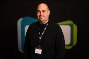 A person with a lanyard and name tag stands in front of a black background with blue and green shapes, representing the distinguished presence of Board Members.