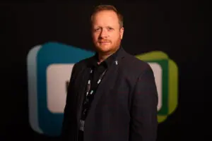 A man with short blond hair and a beard stands indoors among board members, wearing a dark blazer and lanyard, with a blurred abstract background in blue, white, and green tones.