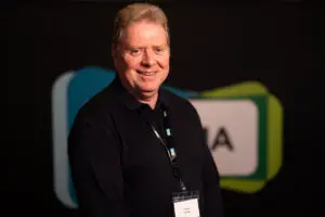 Smiling man in dark shirt with a name badge stands indoors in front of a blurred background featuring a sign with partial letters, suggesting he may be one of the board members at the event.