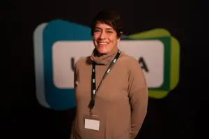 A person wearing a tan polo neck and a lanyard with a blank badge stands smiling in front of a dark background with layered shapes and blurred text, presenting the professional demeanour often associated with Board Members.