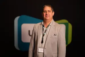 A person in a light grey blazer and lanyard stands in front of a blurred backdrop with blue and green shapes and partial text, presenting a professional image often associated with Board Members.