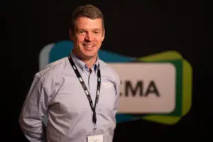 Man wearing a light blue shirt and lanyard stands in front of a blurred sign with partially visible letters MA on a dark background, suggesting he may be one of the board members at the event.