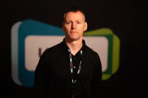 A man in a black shirt and lanyard, possibly one of the board members, stands in front of a blurred background with blue and green shapes.