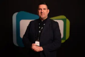 A man wearing a dark blazer and lanyard stands in front of a dark background with overlapping blue and green rectangular shapes, presenting a professional image suitable for Board Members.