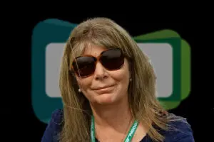 Woman with long hair wearing sunglasses and a lanyard, standing in front of a geometric background with green and blue shapes, representing the UKCMA Fellows community.