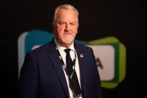 A man in a navy suit and tie stands in front of a dark background with abstract shapes, wearing a lanyard around his neck, representing the distinguished Fellows of the UKCMA.