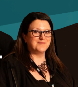 A woman with straight dark hair and glasses wears a black outfit and a lanyard, standing in front of a blue and brown background, presenting the composed professionalism often seen among Board Members.
