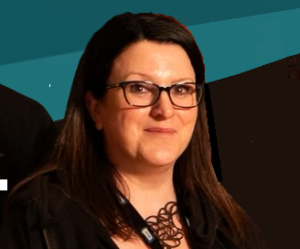 A woman with long brown hair and glasses is facing the camera and smiling slightly, wearing a black top against a teal and black background, presenting a professional image often seen among board members.