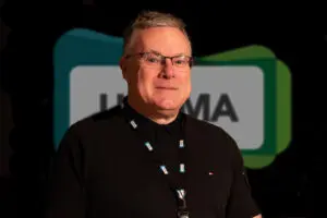 A person wearing glasses and a black shirt with a lanyard, possibly a UKCMA Fellow, stands in front of a blurred sign with partially visible letters.