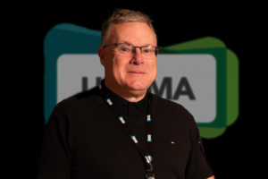 A man wearing glasses and a black shirt with a lanyard, possibly attending a UKCMA event, stands in front of a blurred, colourful background with partially visible text, suggesting his association with other Fellows.