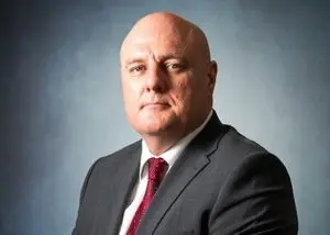 Bald man in a dark suit and red tie poses against a plain, gradient grey-blue background, looking at the camera with a neutral expression, embodying the composed professionalism often seen among board members.