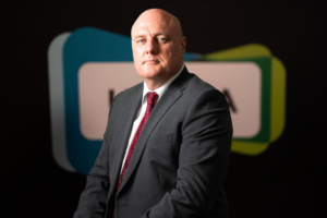A man in a dark suit and red tie poses against a blurred background featuring overlapping shapes, partial text, and a subtle nod to Board Members, suggesting an atmosphere of corporate professionalism.