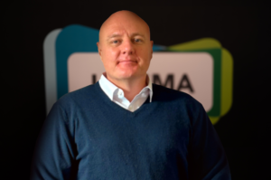 A bald man wearing a blue jumper over a white shirt stands in front of a dark background with partially visible colourful shapes and text, embodying the professional presence often seen among Board Members.