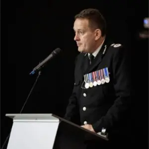 A uniformed man with medals, identified as B. J. Harrington, speaks at a lectern with a microphone, facing left, against a dark background during the QPM 2025 event.
