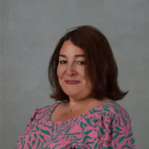 Michelle Russell, a woman with shoulder-length brown hair wearing a pink top with green patterns, poses against a plain grey background in this 2025 portrait.