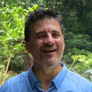 A man with short dark hair and a salt-and-pepper beard smiles outdoors, wearing a light blue shirt. Lush green foliage and sunlight are visible in the background, giving Philip Kolvin, KC an approachable yet distinguished presence.