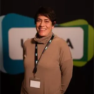 A person with short dark hair wears a beige polo-neck and a lanyard, standing in front of a blurred conference backdrop with indistinct text and colourful shapes, resembling Anne Marie Chebib attending a professional event.
