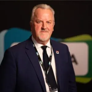 Eric Stuart, dressed in a navy suit and tie with a QPM lanyard, stands in front of a dark backdrop with blurred colourful shapes, confidently looking at the camera as he prepares for the 2025 event.