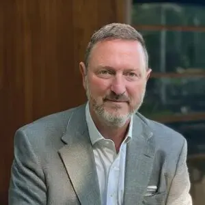 Robert Kaiser, a middle-aged man with short grey hair and a beard, is wearing a grey blazer and white shirt. He is sitting indoors in 2025, with wood panelling and a window in the background.