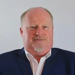 Bald man with short fair hair and goatee wearing a white shirt and dark jacket, facing forwards against a plain light background. Tim Roberts is pictured here in a classic 2025 professional portrait style.