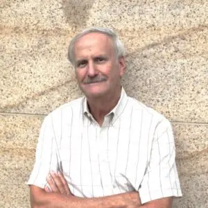 An older man with white hair and a moustache stands against a speckled stone wall, wearing a short-sleeved, light-coloured checked shirt with his arms folded. Reminiscent of Ira Rosen in 2025, he exudes quiet confidence and timeless wisdom.