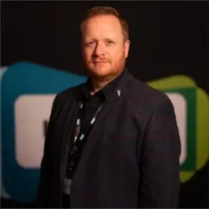 A man with fair hair and a beard wears a dark checked blazer and black shirt, standing in front of a blurred background with geometric shapes—an image that captures Oliver Gardiner’s distinctive style in 2024.
