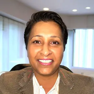 Person with short dark hair and a nose ring, wearing a blazer and white shirt, smiling at the camera in a well-lit office with large windows and recessed lighting. This 2025 portrait captures the confident presence of Satia Rai in a modern professional setting.