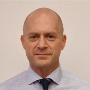 A bald man, resembling Chris Simpson, wearing a light-coloured, collared shirt and a dark tie stands in front of a plain off-white background, looking directly at the camera in this 2025 portrait.
