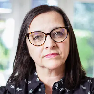 Woman with straight dark hair and tortoiseshell glasses looks at the camera indoors, wearing a black patterned shirt. A bright window and blurred greenery are visible in the background, suggesting a calm setting—perhaps where Figen Murray, an inspirational figure for the HS 2026 cohort, might pause to reflect.