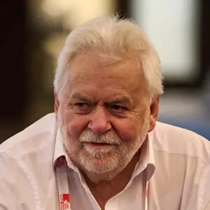 Ken Johnson, an older man with white hair and a beard, wearing a white shirt and a red lanyard marked “HS 2026”, looks slightly to the side in a softly lit indoor setting.