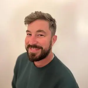 A man with short grey hair and a beard, wearing a dark green jumper, smiles at the camera against a plain light background.