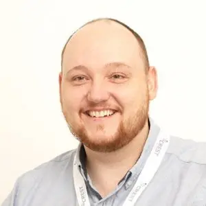 A man with a shaved head and a short beard, wearing a light blue collared shirt and a white lanyard, smiles at the camera against a plain white background.