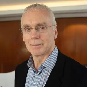 An older man with short grey hair, glasses, and a checked shirt under a black blazer stands in front of a wooden background.