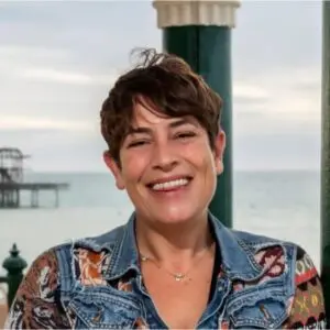 Woman with short brown hair smiling, wearing a patterned shirt and denim jacket, stands outdoors near the seaside with a pier and cloudy sky in the background.